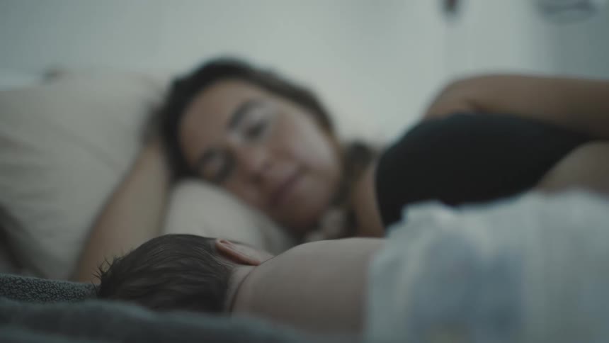 A woman lies in bed watching over a sleeping baby beside her