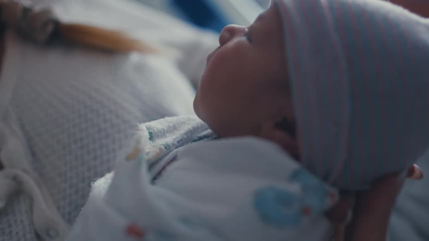A shot focuses on a woman as she smiles and looks at a newborn baby that is not visible on camera she appears to be in a hospital bed holding the baby with a smile on her face
