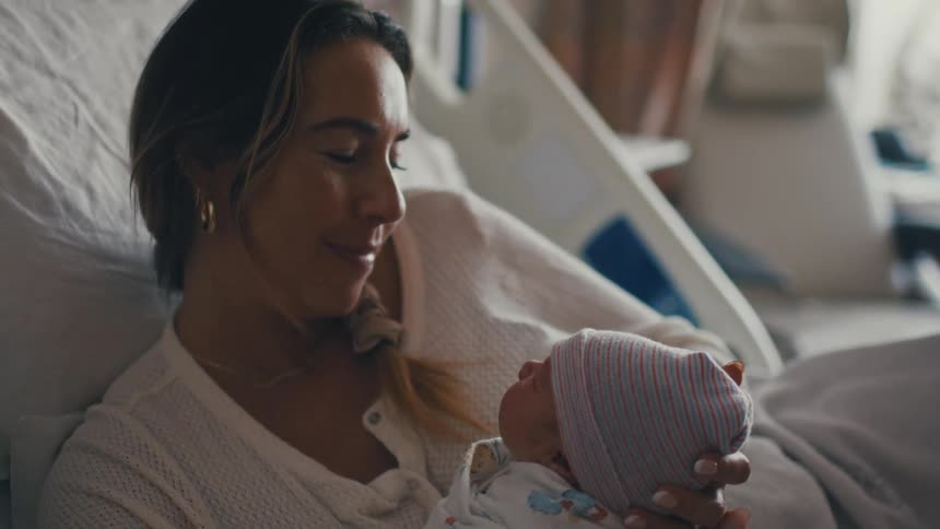 A smiling young woman wearing a white sweater is lying in a hospital bed holding and gently kissing a newborn baby in a striped knit beanie