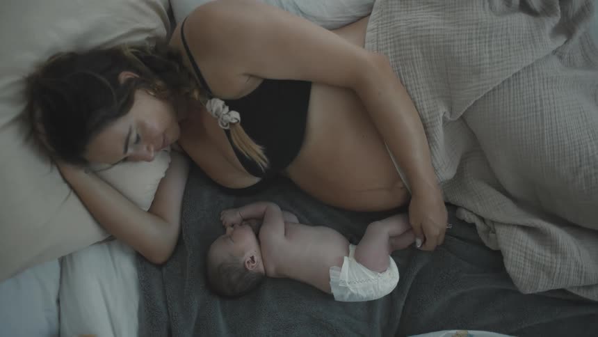 A young woman wearing a bra and covered from the waist down with a blanket lies on the bed smiling as she gazes at her newborn beside her who is wearing only a diaper and sleeping