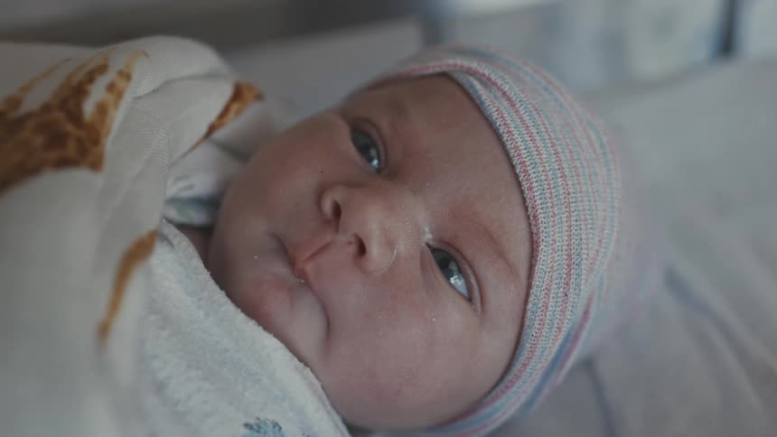 A newborn baby wrapped in a blanket with a striped cap gazes upward with calm curious eyes
