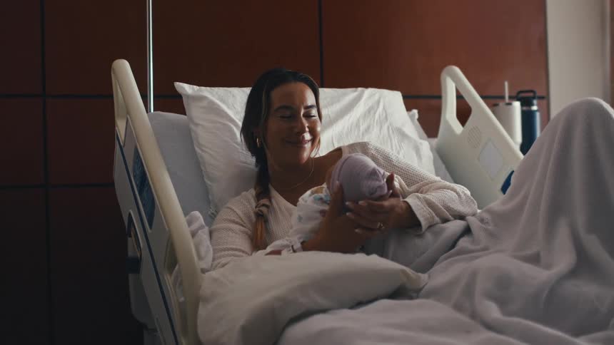 A smiling young woman wearing a white sweater is lying in a hospital bed holding and looking down at a newborn baby wrapped in a swaddle