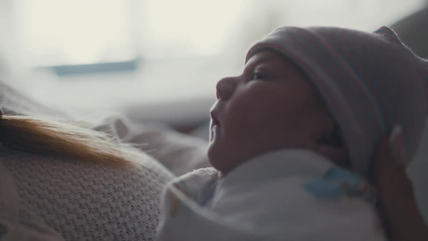 A close view of a smiling young woman lying in a hospital bed holding and looking lovingly at a newborn baby with a knit beanie