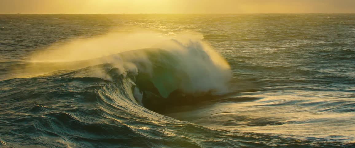Backlit by a golden sunset a powerful ocean wave breaks in slow motion as wind whips spray from its crest