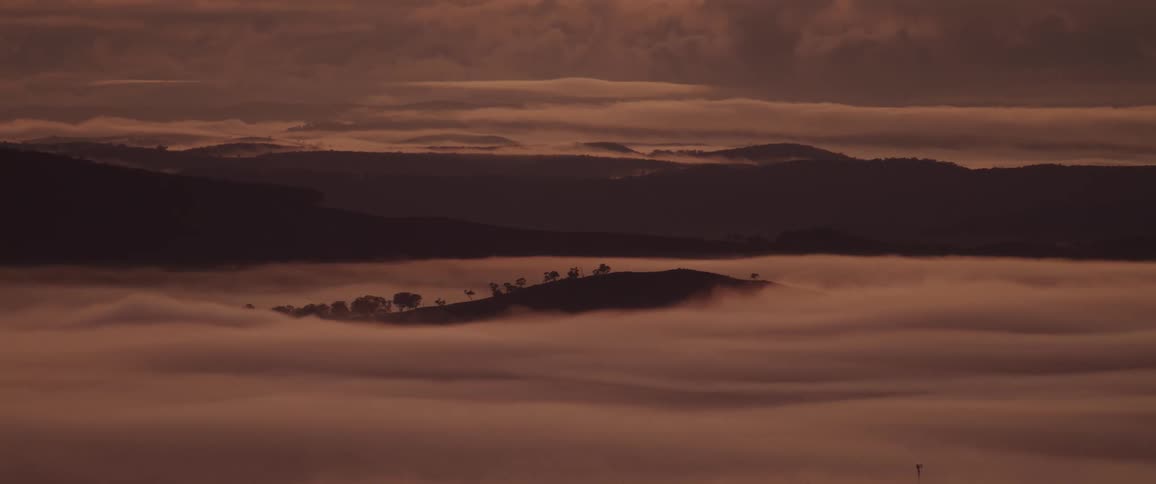 A beautiful natural view of rolling hills and mountains shrouded in a thick layer of fog under a dramatic cloudy sky at sunset
