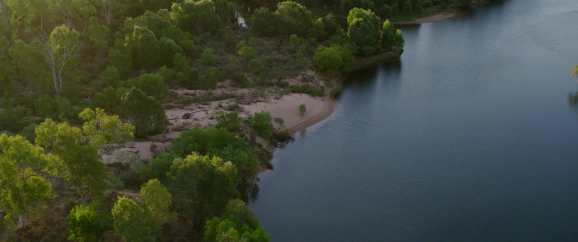 A calm river curves beside a sandy shoreline and dense green forest in the warm evening light