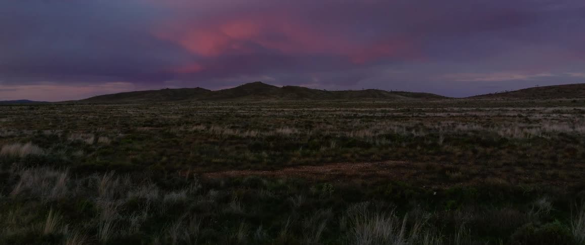 A vast dry landscape of grass and shrubland stretches out toward a distant mountain range under a dramatic evening sky with swirling dark and reddish clouds