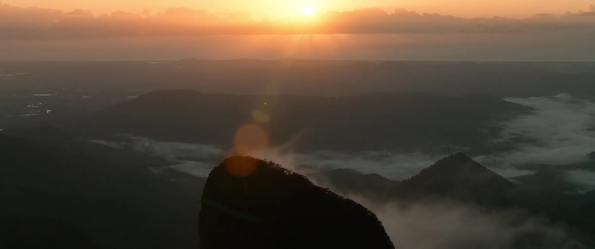 An orange sunset casts its glow over the wild as foggy white clouds drift around the rocky mountainous terrain