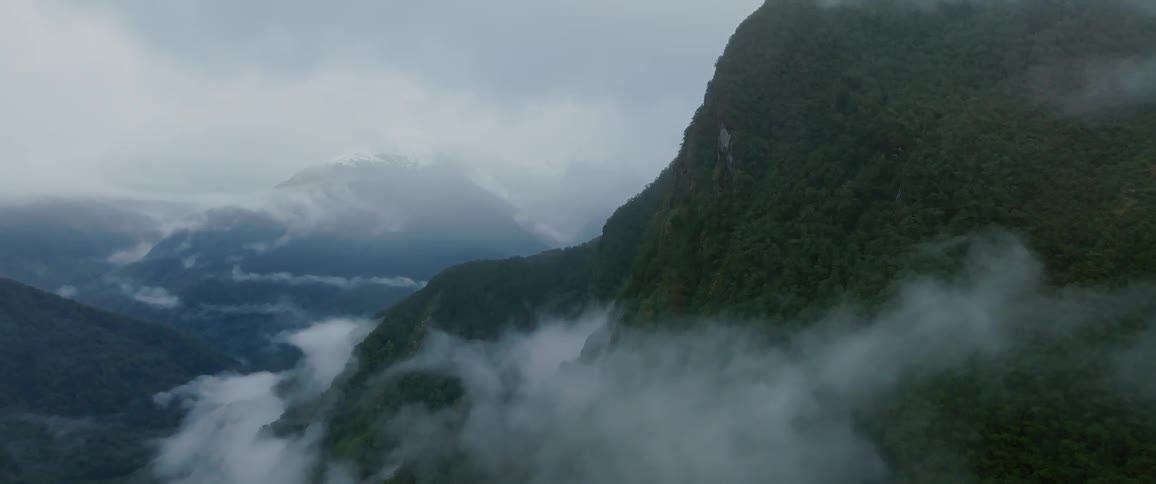 A mountainous view features a peak covered in green vegetation surrounded by a foggy atmosphere at midday