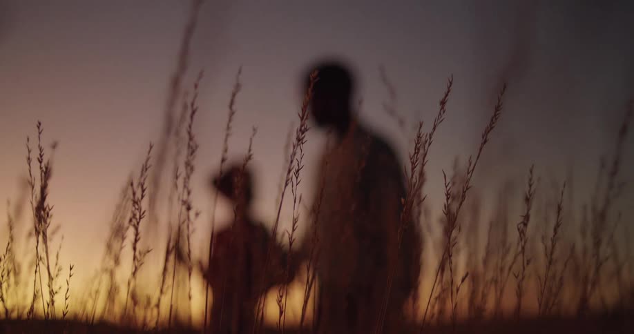 An adult and a child stand in a field of tall grasses at sunset