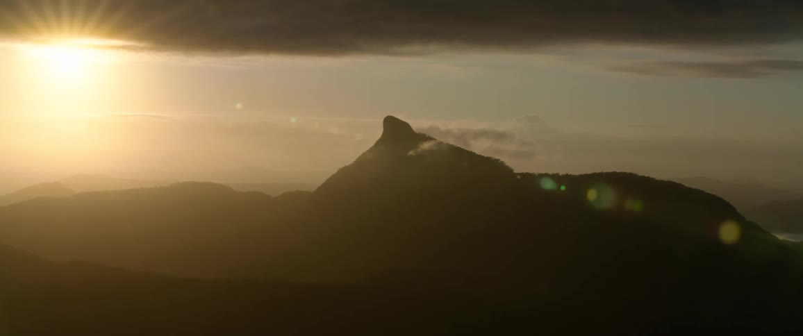 A low aerial shot shows a rugged pointed mountain peak silhouetted against a cloudy sky during a hazy sunset with the last light of the day casting a warm glow and lens flare