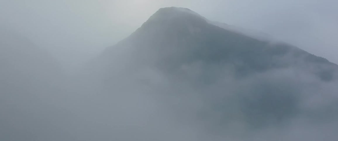 A wide view shows the peak of a dark rocky mountain shrouded in thick fastmoving clouds and fog creating a moody and isolated atmosphere