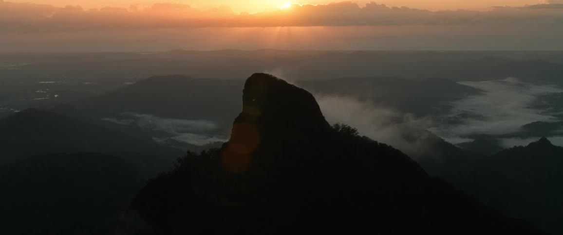 Sunbeams from a golden sunrise break over a silhouetted mountain peak with low lying clouds filling the hazy valleys