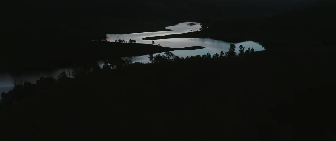 A dark wide aerial view shows a winding river in a remote mountainous landscape with trees silhouetted against the waters surface as a bright reflection fades in the gloomy atmosphere