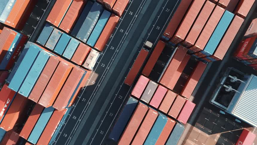 A container truck drives through a seaport on a tarred road with stacked cargo containers towering on both sides at midday