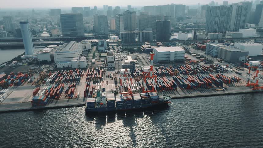 An industrial port with numerous shipping containers cranes and a large cargo ship is bustling with activity set against a city skyline