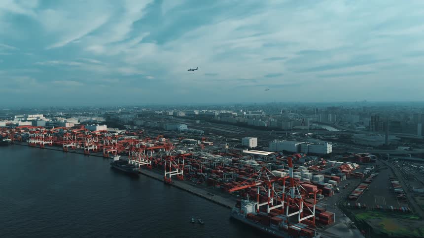 An expansive aerial view captures a sprawling industrial port filled with container ships and cranes extending towards a distant city skyline under a cloudy sky with airplanes flying overhead