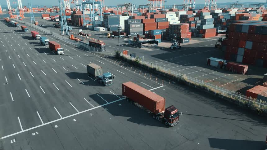 Trucks loaded with containers drive out of a busy seaport at midday while empty trucks head in passing towering stacks of colorful cargo containers under the bright sun