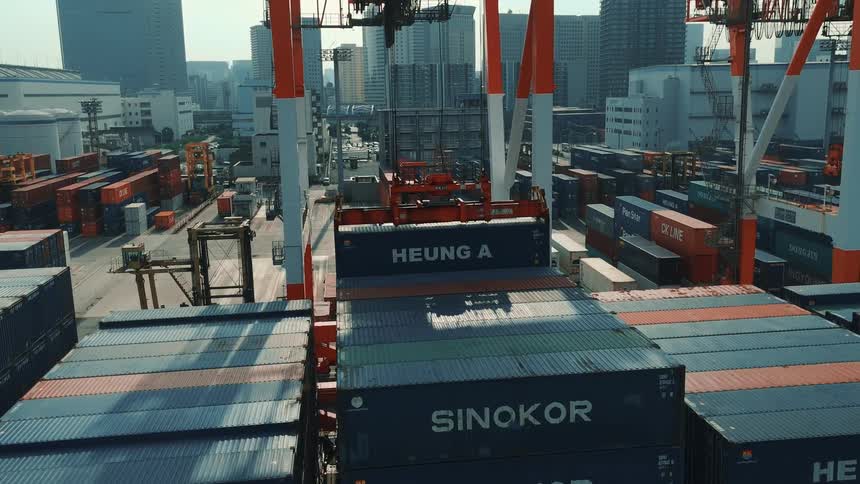 Crane moves blue containers in a cargo area surrounded by urban buildings