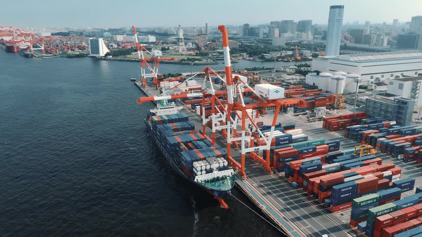 This aerial video shows a cargo ship at a busy industrial port with many colorful containers stacked on the dock large cranes are visible and in the background you can see a city skyline with various buildings and structures
