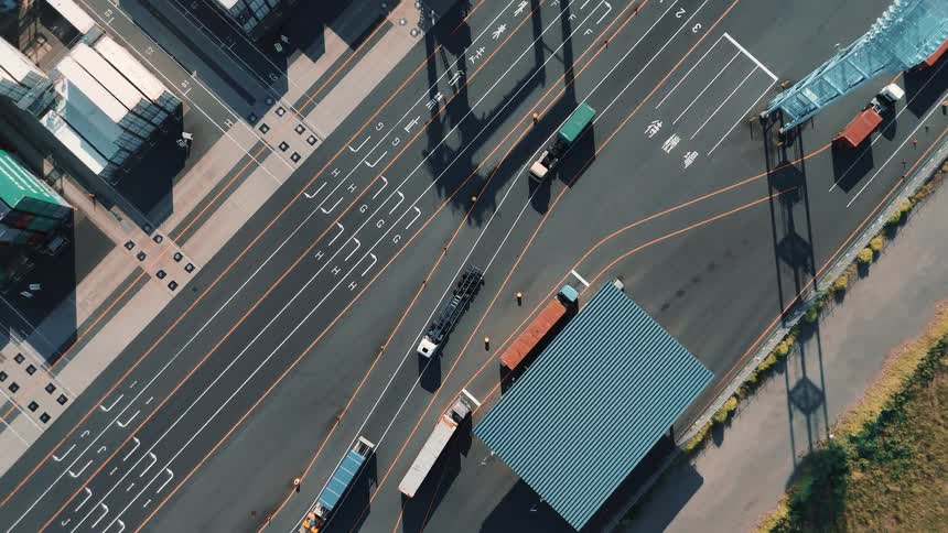 Trucks and shipping containers move through a port terminal seen from above with marked lanes and storage areas