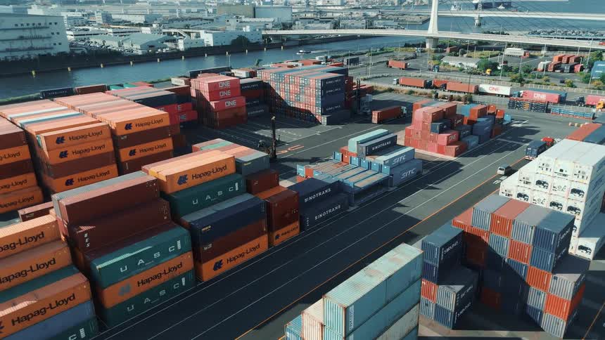 This drone footage captures an aerial view of a container port where rows of stacked shipping containers are organized in the background a bridge is visible over a body of water and various industrial buildings and vehicles are scattered around