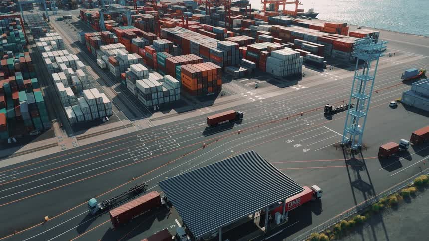 An aerial view of a busy industrial port shows numerous shipping containers of various colors stacked high with trucks moving along the pavement and large cranes visible in the background against the bright sunlight reflecting on the sea
