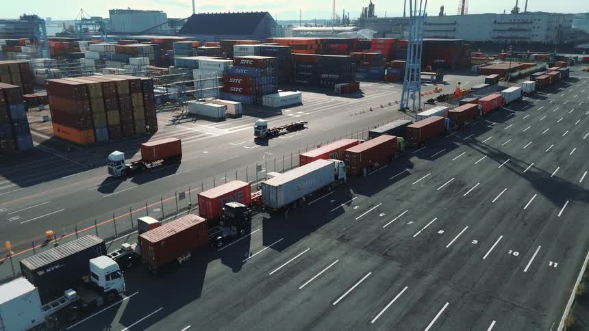 A seaport at midday with a road lined with cargo trucks flanked by rows of stacked shipping containers all set against the backdrop of surrounding ocean waters