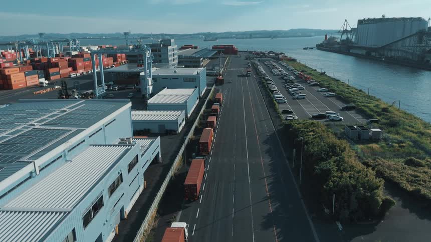 A seaport at midday with a road lined with cargo trucks flanked by rows of stacked shipping containers all set against the backdrop of surrounding ocean waters