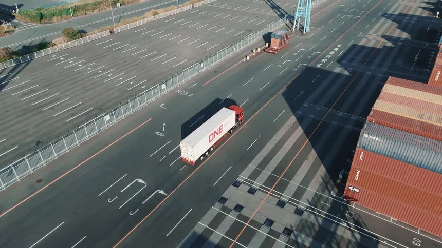The video shows an aerial view of a busy shipping yard with two distinct lines of stationary objects the first line is composed of several rows of stacked shipping containers these containers are varied in color with many being blue orange or red