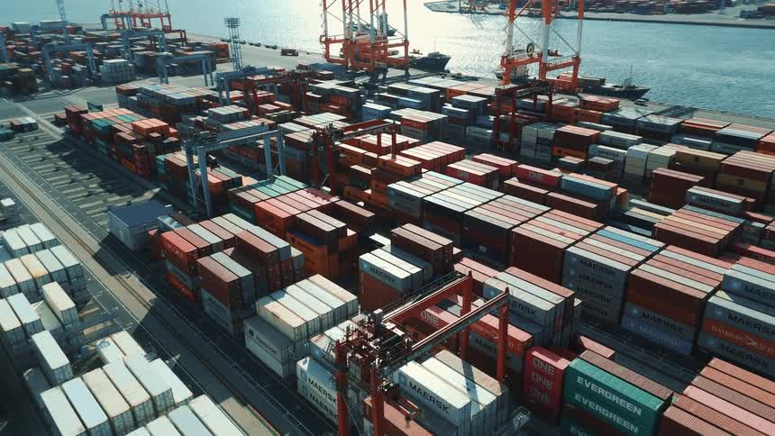 A vast shipping port filled with stacked containers stretches along the waterfront with cranes and ships actively loading and unloading cargo