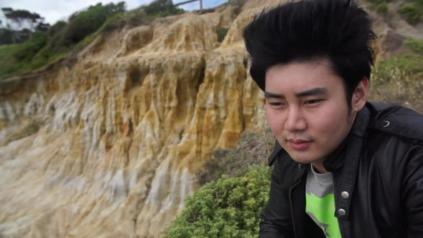 The wind blowing a young, asian man's hair straight up as he sits near brush and a cliff