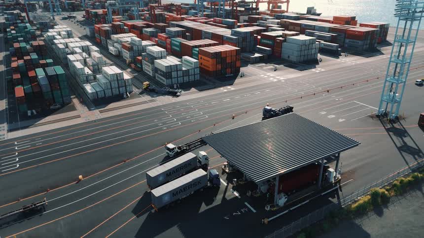 A series of trucks drive into a seaport at midday heading along paved lanes between towering stacks of cargo containers ready to pick up shipments under the bright sun