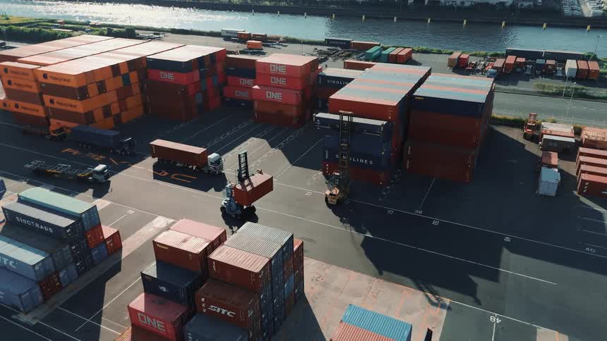 This is an aerial video of a busy shipping yard the footage shows a forklift carrying a shipping container with many other containers stacked in the background a bridge and a waterway are also visible in the distance