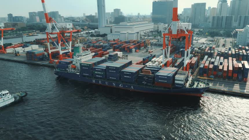 An aerial shot captures a large cargo ship filled with containers docked at a busy port in the background a modern city skyline rises behind the towering cranes used for loading and unloading