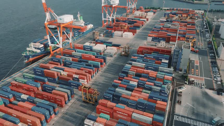 Stacks of colorful shipping containers fill a busy port terminal with cranes and trucks operating near the water
