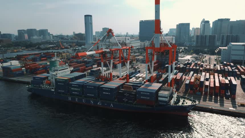 A cargo ship is docked in a busy sea port harbor at midday with two towering cranes hovering above as they work the vessel the ships deck is stacked high with colorful shipping containers while more containers are piled in the background on the port
