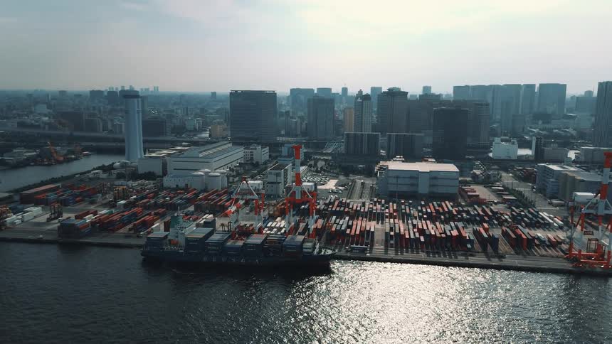 A bustling seaport at midday filled with stacks of colorful cargo containers in the harbor a cargo ship loaded with containers sits docked while a towering crane hovers above the city skyline rises in the background under the bright sun