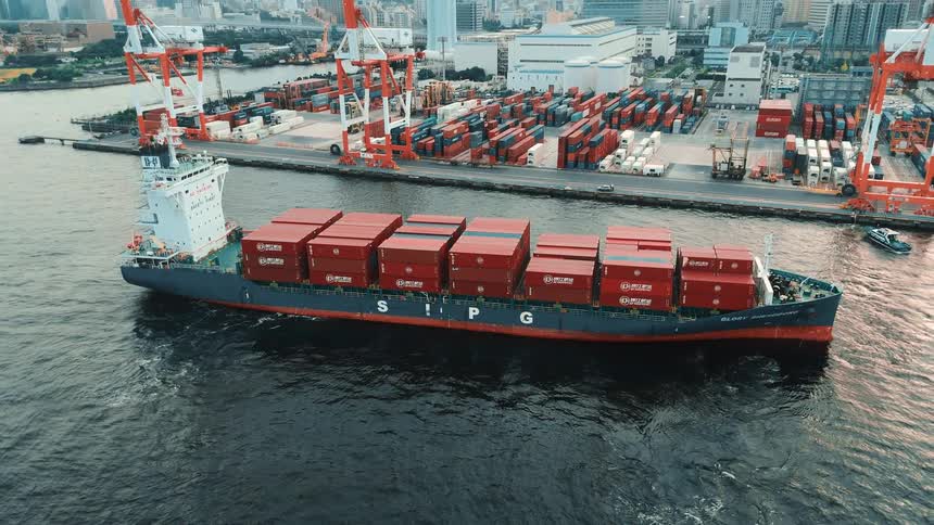 Freight ship full of containers on the water next to a port with a metropolitan area in the background outdoors during day time