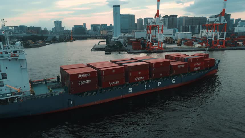 A boat loaded with cargo containers approaching a commercial port with a large shipping container depot