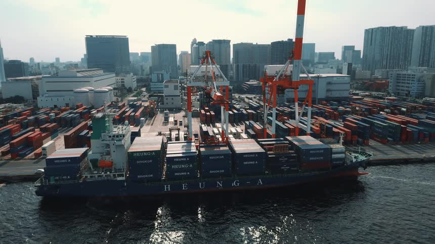 A seaport at midday with a cargo ship docked in the harbor its deck loaded with rows of stacked containers towering cranes stand beside the vessel poised for loading and unloading while more stacks of containers fill the background across the port