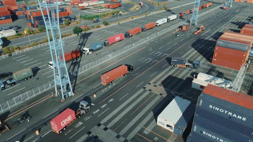 An aerial drone view shows many trucks moving cargo containers around a large port on a sunny day