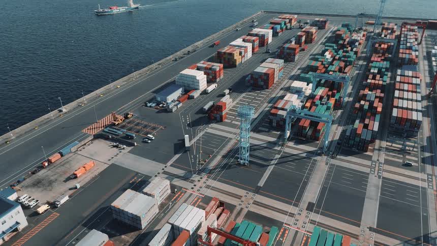 A commercial port with a large cargo shipping container depot with a boat approaching and cargo trucks moving inside it