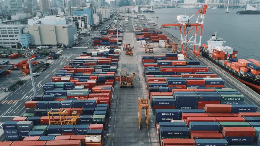 A wide view of a busy port shows countless shipping containers lined up along the waterfront with cranes and city skyscrapers in the distance