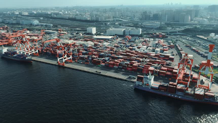 A seaport at midday with two cargo ships docked in the harbor their decks stacked high with containers massive cranes tower overhead while trucks drive through the port past rows of stacked containers with the city skyline visible in the background