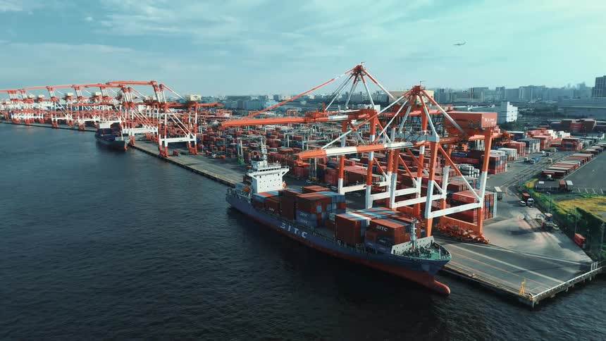 Cargo ship full of shipping containers docked on a port next to a metropolitan area outdoors during day time