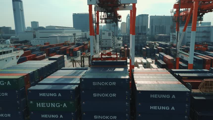 This is an aerial video of a bustling seaport on a bright day the camera pans over a cargo ship showing towering cranes actively moving and arranging large shipping containers on the vessel and the surrounding docks with a city skyline visi