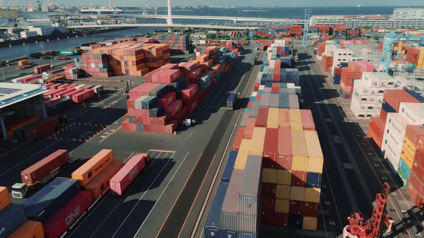 A seaport at midday with rows of stacked cargo containers as trucks drive by carrying containers along the paved lanes