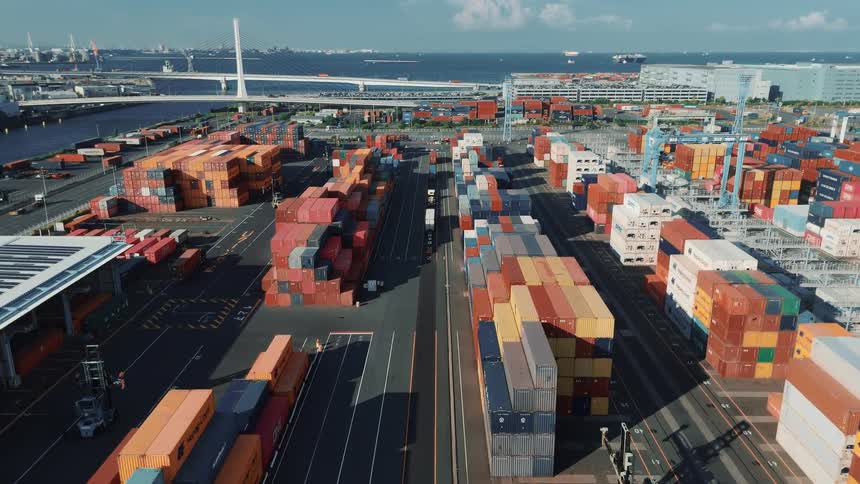 A busy seaport at midday packed with towering stacks of colorful cargo containers as trucks loaded with container freight drive in and out along the paved lanes under the bright sun