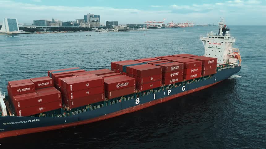 This drone video captures an aerial view of a large cargo ship as it sails on the water the ship is loaded with numerous red shipping containers and is sailing with a large port and city skyline in the background